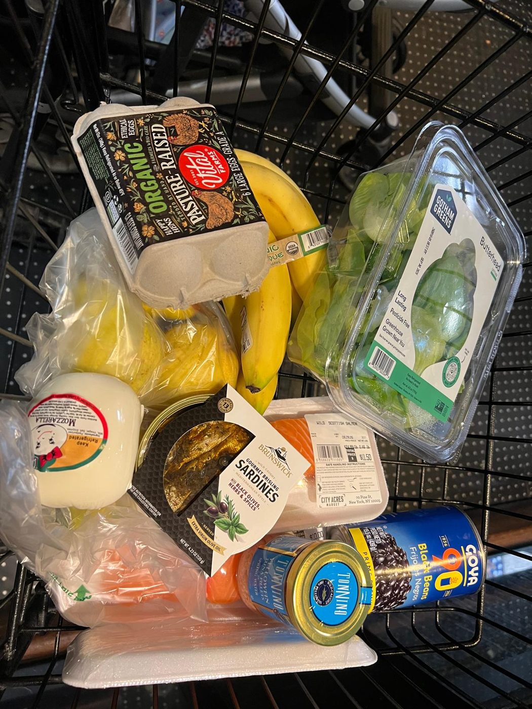 A shopping cart with eggs, bananas, greens, mozzarella, sardines, tuna and black beans