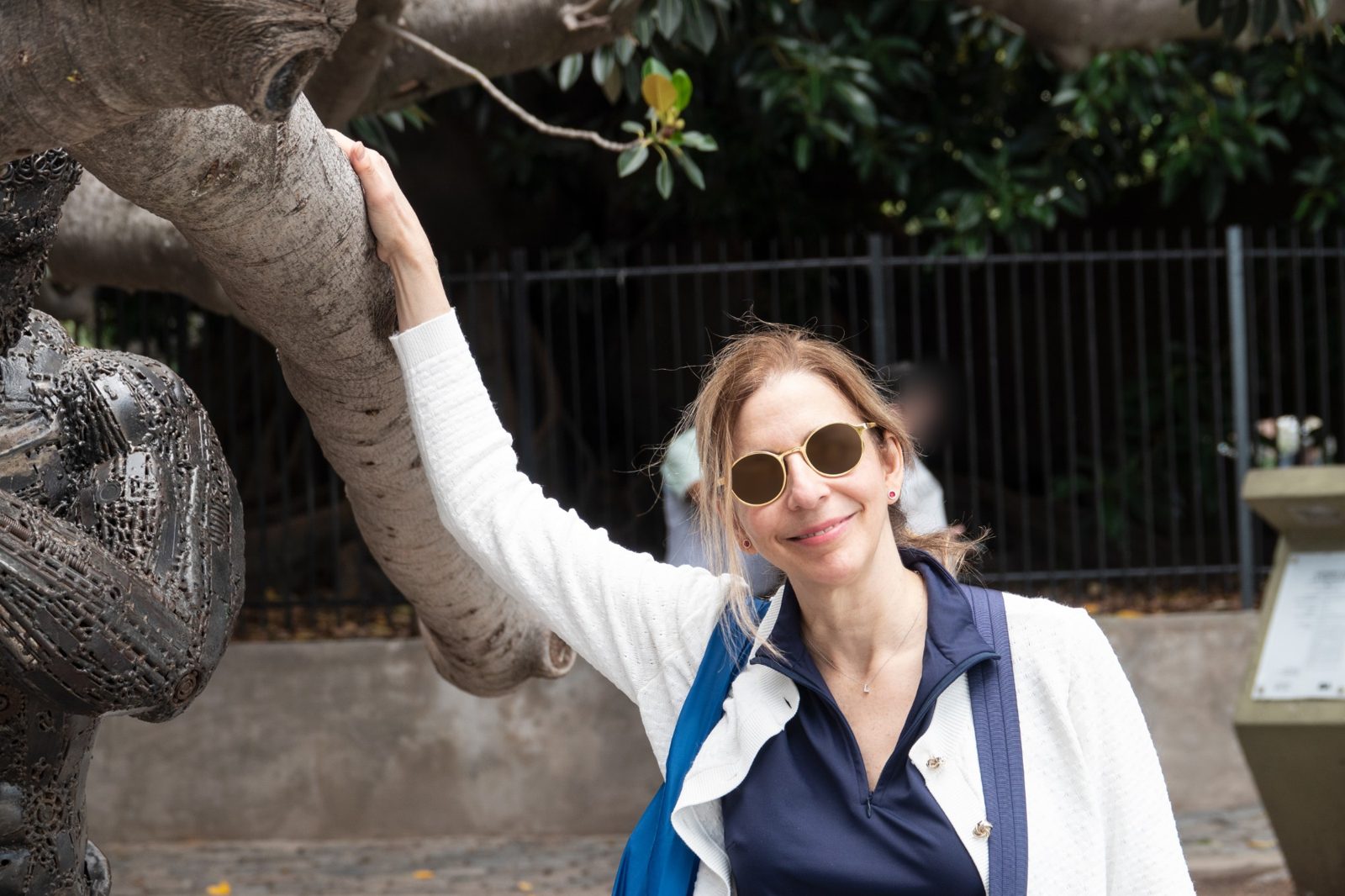 Linda Purpura Fischer in sunglasses, one hand resting on a tree branch beside a metal sculpture